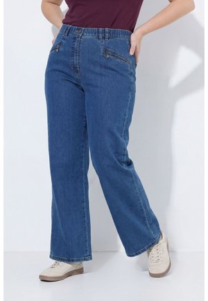 Frau trägt blaue Jeans mit weitem Bein und Reißverschlusstaschen vorne sowie beige Sneaker, steht vor einem einfarbigen weißen Hintergrund.