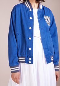 Modrá bunda s předním zapínáním na knoflíky, bílými knoflíky a nášivkou ve tvaru štítu. Má pruhované manžety a límec.