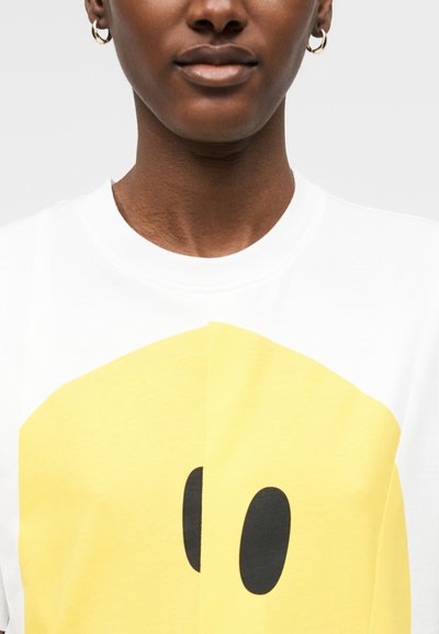Personne portant un t-shirt blanc avec un grand smiley jaune et des yeux ovales noirs, boucles d'oreilles en anneau doré, fond neutre.