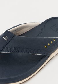 Mørkeblå flip-flop med strukturert overdel, hvit aksentlinje, kremfarget såle og gult "REEF"-logo detalj.