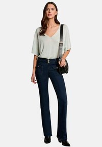 Top verde claro de manga corta y escote en V combinado con jeans acampanados de cintura alta en azul oscuro, con bolsillos frontales y detalles de botones. Bolso negro.