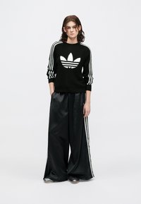 Suéter preto de malha com detalhes em branco em três riscas e grande logótipo trefoil branco, combinado com calças largas pretas brilhantes e sapatos prateados.