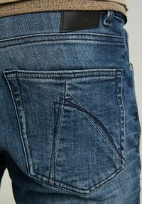 Jean en denim bleu avec une poche arrière détaillée arborant un motif de couture triangulaire unique, accompagnée d'une étiquette en cuir noire texturée.