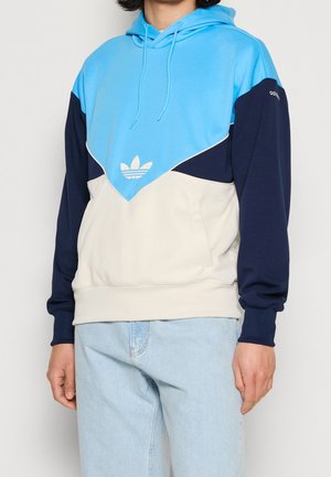 Mężczyzna w bluzie z kapturem w kolorach niebieskim, granatowym i beżowym z logo Adidas na piersi i rękawie, w połączeniu z jasnoniebieskimi dżinsami.