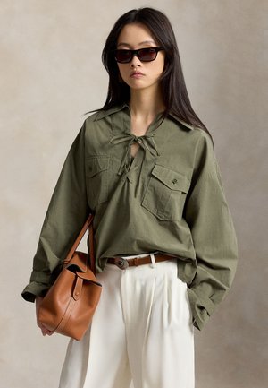 Donna che indossa una camicia verde oliva oversize con collo allacciato, pantaloni bianchi plissettati, occhiali da sole scuri e porta una borsa in pelle marrone.