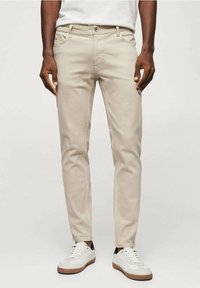 Jeans slim fit beige con bolsillos frontales y cierre de botón, fabricados en denim de algodón. Combinados con zapatillas blancas con suelas marrones.