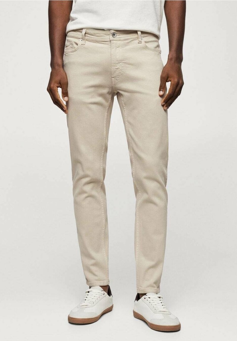 Jeans slim fit beige con bolsillos frontales y cierre de botón, fabricados en denim de algodón. Combinados con zapatillas blancas con suelas marrones.