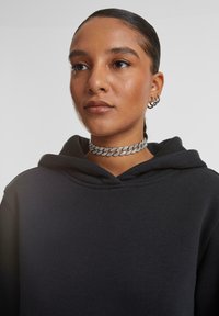 Femeie cu păr neted tras înapoi poartă un choker din lanț argintiu, cercei de tip cerc și un hanorac negru, privind ușor în lateral.