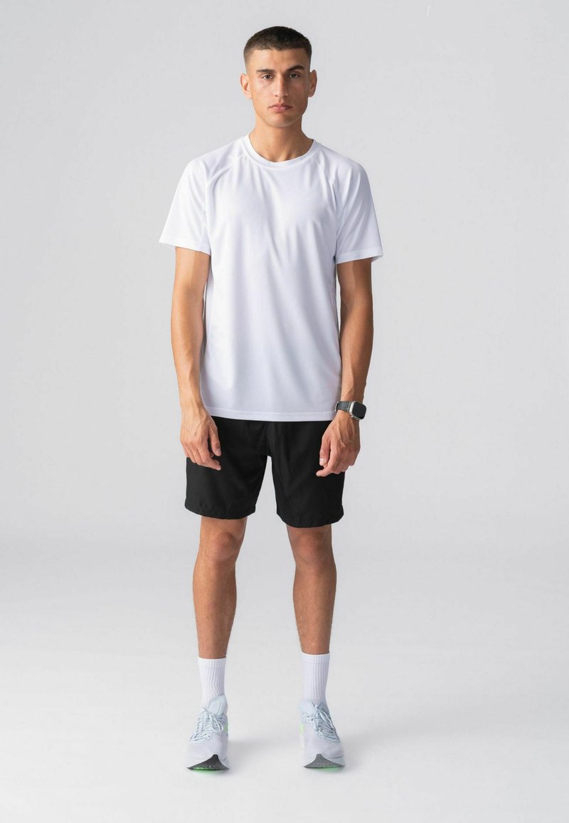 Jeune homme debout face à l’avant, portant une chemise blanche à manches courtes, un short noir, des chaussettes blanches, des baskets claires et une montre connectée noire.