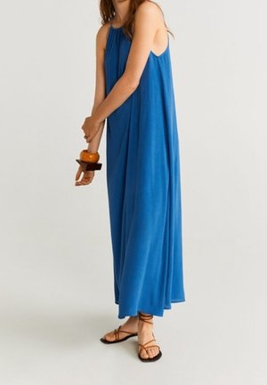 Femme portant une robe longue bleue sans manches et ample, avec des sandales marron à lanières et de grands bracelets en bois au poignet gauche.