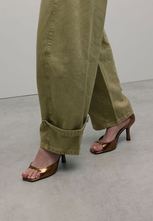 Sandales à talons hauts en bronze avec un design à bout ouvert et une texture brillante, associées à un pantalon large vert olive clair, retroussé aux chevilles.