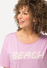 Lyserød t-shirt med hvid, fluffy "BEACH" skrift. Korte ærmer og rund halsudskæring med en blød, glat tekstur.