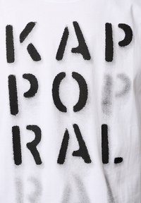 T-shirt en coton blanc avec de grandes lettres noires en gras épelant "KAPORAL". Le texte semble légèrement usé avec un aspect texturé.