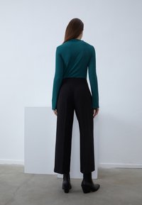 Inimene, kellel on pikk juuksed, kannab türkiissinist pika varrukaga särki, mustad laiade säärega pükse ja musti pahkluu saapaid, seisab siseruumis valge seina ees.