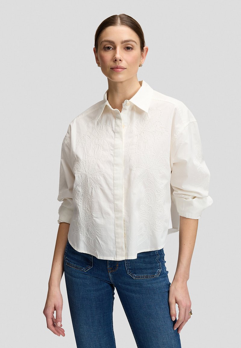 Femme aux cheveux bruns portant une chemise blanche brodée à manches longues et un jean bleu, debout devant un fond gris uni.