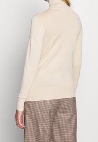 Kvinna i en krämfärgad polotröja och bruna rutiga vida byxor, sedd bakifrån mot en enkel grå bakgrund.