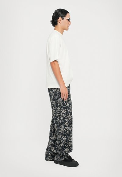 Weißes Kurzarm-T-Shirt kombiniert mit weiten schwarzen Blumenmuster-Hosen und schwarzen Sneakers. Das Model steht seitlich und präsentiert das Outfit.