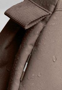 Tessuto impermeabile marrone con superficie testurizzata, caratterizzato da gocce d'acqua. Include una patta pieghevole e un dettaglio con cursore in metallo.