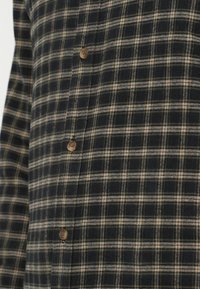 Πουκάμισο flannel σε ναυτικό και μπεζ καρό σχέδιο, με κουμπωτές κλείσεις και απαλή υφή. Το ύφασμα φαίνεται ζεστό και ανθεκτικό.