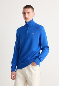 Blauer Strickpullover mit hohem Kragen und Viertelreißverschluss, ausgestattet mit gerippten Manschetten und Saum, kombiniert mit hellen Hosen. Goldenes Logo-Detail sichtbar.