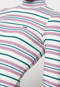 Haut à manches longues côtelé avec des rayures verticales en lavande, blanc et sarcelle. Col montant avec un logo subtil "Lee" en noir.