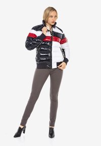 Wattierte Jacke in Marineblau, mit weißen und roten Streifen; glänzendes Finish; hoher Kragen; Frontreißverschluss; gerippte Bündchen; kombiniert mit dunklen Hosen und High Heels.