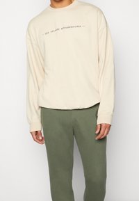 Beige Sweatshirt mit langen Ärmeln und dem Aufdruck „LES VOYAGES EXTRAORDINAIRES“ auf der Vorderseite. Kombiniert mit olivgrünen Jogginghosen.