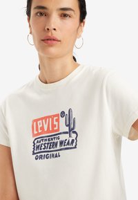 Valge puuvillane T-särk, millel on graafiline trükk punases, sinises ja mustas, millel on kirjad "Levi's Autentne Lääne Riietus" ning kaktuse kujundus.