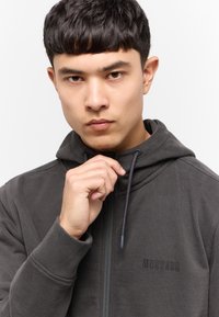 Sudadera con cremallera de color gris oscuro, con una superficie texturizada, cordón ajustable y logotipo bordado de "MUSTANG" en el lado izquierdo.