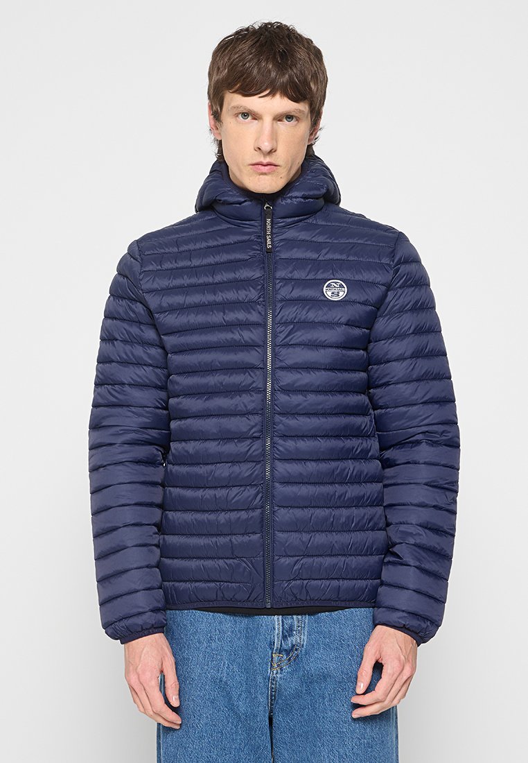 Mężczyzna w granatowej pikowanej kurtce puchowej z kapturem i zamkiem, w połączeniu z niebieskimi dżinsami, stojący na jednolitym tle.