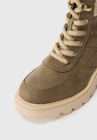 Cizme scurte din suede verde măsliniu, cu șireturi bej, căptușeală pufoasă și o talpă chunky bej cu un model texturat al benzii de rulare.