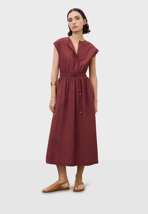 Mujer con vestido midi sin mangas color granate con cintura con cordón, sandalias planas marrones, pulseras doradas y pendientes de aro sobre fondo liso.