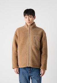 Brun fleecejakke med lynlås, høj krave, sidelommer og en brystlomme med en stofdetalje. Casual design og tekstur.