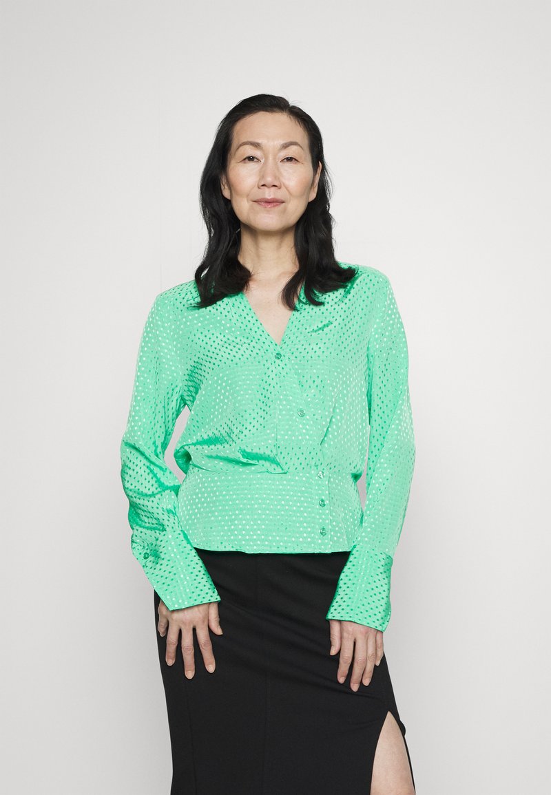 Blusa verde menta com botões, feita de material leve, apresentando um padrão de bolinhas, mangas longas e decote em V, combinada com uma saia preta.