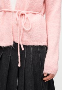 Μαλακή ροζ πλεκτή ζακέτα δεμένη στη μέση πάνω από λευκό πουκάμισο, σε συνδυασμό με σκούρα πλισέ φούστα, με το χέρι να ακουμπάει στο πλάι.