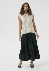 Vaalean kultainen hihaton neulepaita, jossa on teksturoitu kuvio, yhdistettynä mustaan laskostettuun hameeseen ja raidallisiin korkokenkiin.