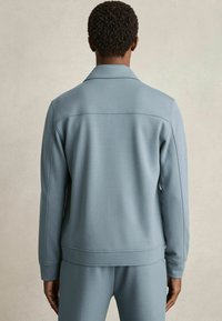 Un hanorac moale, de culoare albastru deschis, cu guler și manșete ribbate, având un design curat și o textură netedă, văzut din spate.