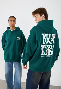 Grüne, oversized Kapuzenjacke mit einer Fronttasche, die auf der Rückseite weißen grafischen Text aufweist und eine Kordelkapuze hat. Getragen mit hellblauen und dunklen Jeans.