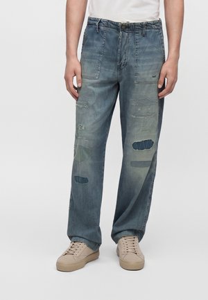 Mand iført løstsiddende blå cowboybukser med flere lapper og beige sneakers, stående mod en ensfarvet baggrund.