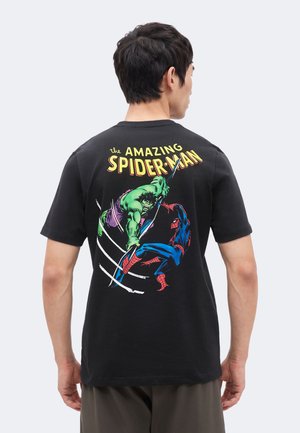 Mies, joka pukeutunut mustaan t-paitaan, jossa värikäs kuva Hulkista ja Spider-Manista taistelemassa tekstin "The Amazing Spider-Man" alla takana.