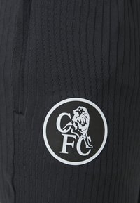 Melnas sporta bikses ar teksturētām svītrām un apaļu baltu logotipu, kurā ir lauva un "CFC", uz apakšējās kājas.