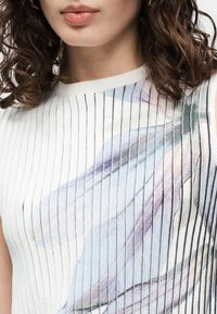 Γυναίκα με καστανά σγουρά μαλλιά που φορά λευκό αμάνικο τοπ με λεπτές μαύρες ρίγες και απαλό παστέλ αφηρημένο μοτίβο.