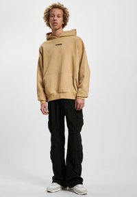 Sudadera beige con bolsillo frontal, pantalones cargo negros con bolsillos laterales y zapatillas blancas. Diseño sencillo, corte relajado y texturas suaves.