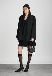 Musthave musta üleriide, topelt rinnavööt, koos musta mini seelikuga, läbipaistvate mustade reisiüleste sukkade ja terava musta kontsakingaga. Väike tekstureeritud käekott.