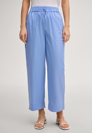 Kobieta ubrana w luźne, niebieskie spodnie o szerokich nogawkach z elastycznym pasem i sznurkiem, w połączeniu z białymi sandałami na paski, stojąca na jednolitym tle.