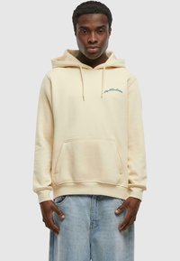 Sudadera color crema hecha de un material suave, con un bolsillo delantero y capucha, y texto bordado en verde en el área del pecho izquierdo.