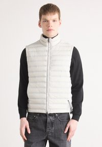 Vadderad ljusgrå puffer-väst med hög krage, dragkedja fram och sidofickor, som bärs över en svart långärmad tröja och grå jeans.