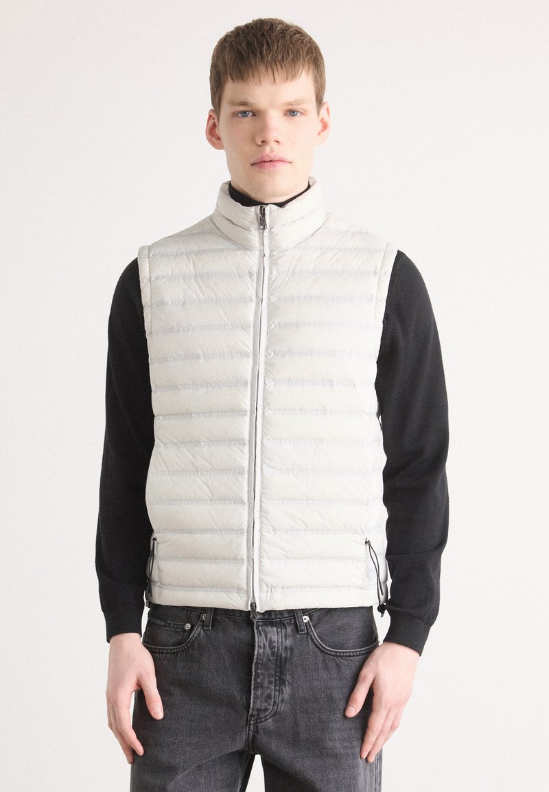 Vadderad ljusgrå puffer-väst med hög krage, dragkedja fram och sidofickor, som bärs över en svart långärmad tröja och grå jeans.