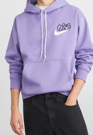 Osoba nosząca lawendową bluzę z kapturem z białymi sznurkami i graficznym logo Nike, połączoną z czarnymi spodniami, jedną ręką w kieszeni.