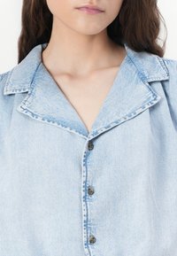 Ανοιχτό μπλε τζιν πουκάμισο με φαρδύ γιακά, μπροστινό κούμπωμα και διακριτή υφή. Εντυπωσιακές λεπτομέρειες είναι οι αντίθετες ραφές και τα σκούρα κουμπιά.